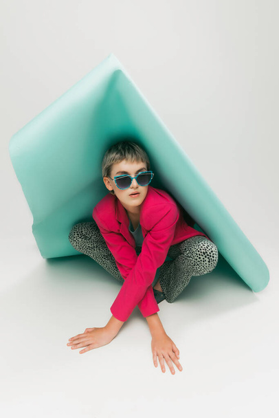 Portrait of young girl in bright jacket and vintage pants posing with paper piece isolated over grey studio background. Concept of retro fashion, art photography, style, queer, beauty - Photo, image