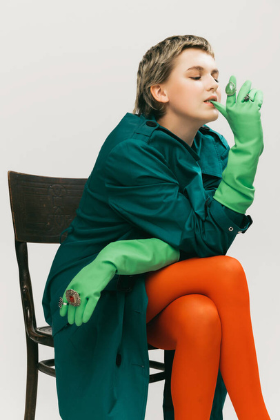 Πορτρέτο της νεαρής κοπέλας σε πράσινο παλτό, πορτοκαλί καλσόν και γάντια από καουτσούκ, κάθεται στην καρέκλα, θέτοντας απομονωμένη πάνω από γκρι φόντο στούντιο. Αντίληψη της ρετρό μόδας, φωτογραφία τέχνης, στυλ, queer, ομορφιά - Φωτογραφία, εικόνα