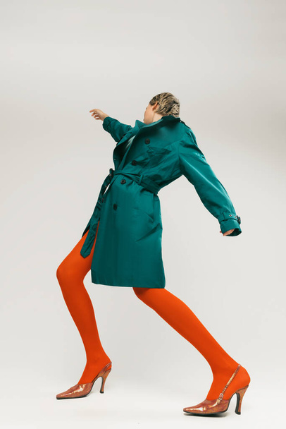 Портрет стильной девушки в зеленом пальто и ярко-оранжевых колготках, танцующих изолированно на сером фоне. Экспрессивная мода. Ретро-мода, арт-фотография, стиль, квеер, красота - Фото, изображение