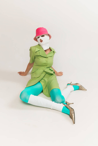Retrato de niña en pasamontañas blanco, panama rosa y traje verde brillante posando aislado sobre fondo gris estudio. Concepto de moda retro, fotografía de arte, estilo, queer, belleza - Foto, Imagen