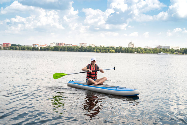 beautiful young woman in a protective vest learns to swim on a sup board on a city lake, active lifestyle, summer time - Photo, Image