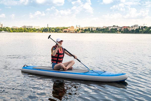 Mujer joven sentada en el tablero de paddle usa chaleco salvavidas y aprende a nadar, estilo de vida activo de verano - Foto, Imagen