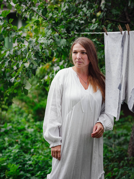 中年白人女性白衣を着た白人女性が宿題をして村のコテージハウスの中庭の路上で服をぶら下げて洗濯日のコンセプト - 写真・画像