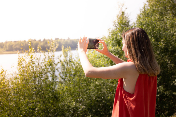 The pretty young caucasian woman in red skirt is taking taking photo by mobile phone in the green forest lakeside at the outside with flare. High quality photo - Photo, Image