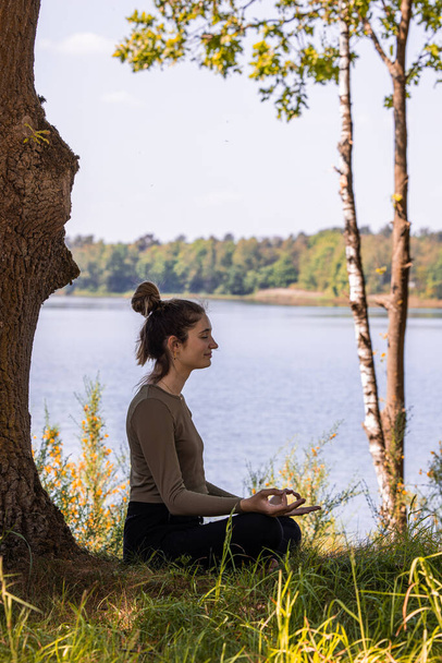 Detailní záběry mladé ženy praktikující jógu v přírodě. Sedni si na zelenou trávu vedle stromu a lesního jezera. Komunikovat pro cvičení a udržovat dobré zdraví. Kvalitní fotografie - Fotografie, Obrázek
