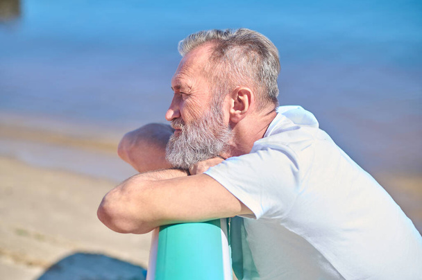 Reflections. Profile of gray-haired bearded man touching surfboard looking thoughtfully on seashore at sunny day - Photo, image
