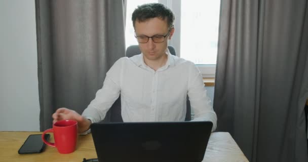 Mladý muž pracuje v laptopu a pije kávu z poháru. Zrychlený pohyb pracovních toků. Koncept vzdálené práce doma, pracovní den, domácí kancelář, dálkové učení, psaní na klávesnici. Portrét, vepředu - Záběry, video