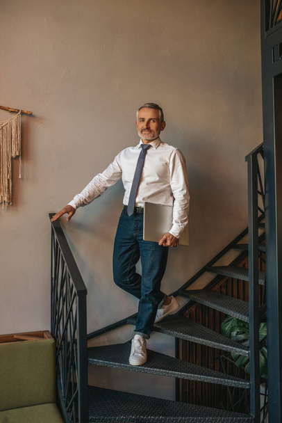 Confidence. Stylish gray-haired man in tie with laptop standing indoors on stairs touching railing looking confidently at camera - Valokuva, kuva