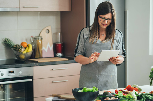 Szczęśliwa kobieta za pomocą tabletu cyfrowego do oglądania samouczków internetowych i podejmowania zdrowego posiłku stojąc przed stołem pełnym świeżych warzyw organicznych w kuchni - Zdjęcie, obraz