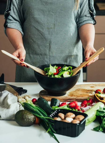 Een anonieme vrouw die gezonde lunch maakt, biologische groenten mengt met een lepel terwijl ze aan het keukenbureau staat met verse ingrediënten - Foto, afbeelding