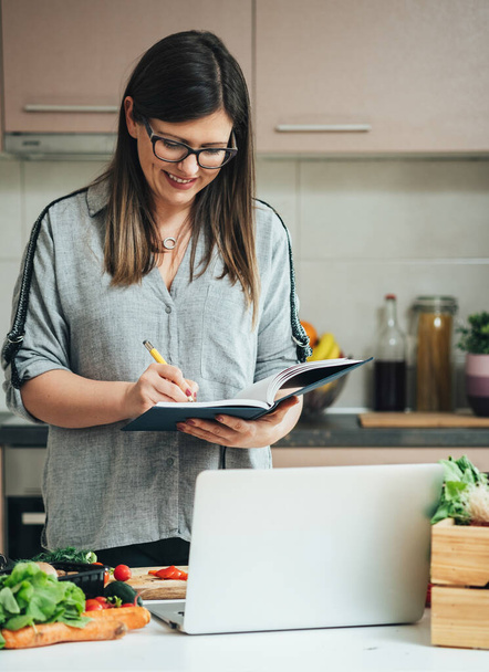 Boldog nő írásban recept egy szakácskönyv és nézi főzés leckék a számítógépen, miközben áll az asztalnál tele friss színes zöldségek házi ebéd - Fotó, kép