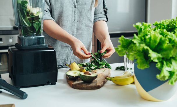 Tanınmayan Kadın Sağlıklı Detoks İçeceğini Karıştırıcı 'da Hazırlıyor: Mutfak masasında dururken muz, ıspanak ve avokado ile yeşil smoothie - Fotoğraf, Görsel