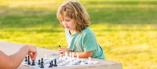 Padre e figlio giocano a scacchi all'aperto, banner poster con spazio copia, figlio bambino che gioca a scacchi sul tavolo nel parco all'aperto con suo padre, infanzia. - Foto, immagini
