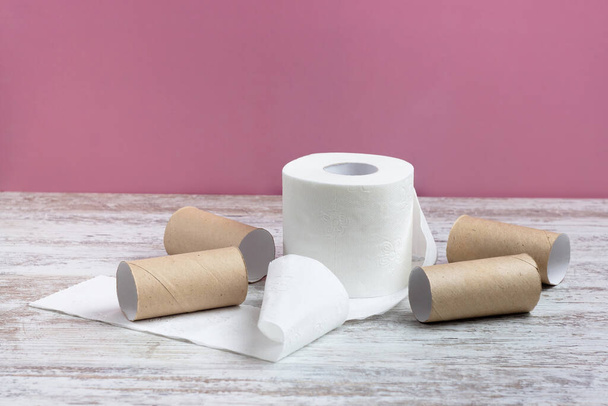 An unwound roll of soft white toilet paper among empty toilet paper rolls. On a wooden table on a pink background - Foto, imagen