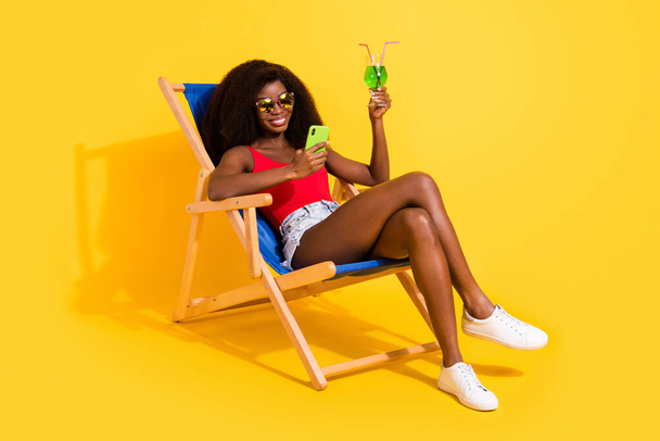 Portrait of attractive cheerful girl drinkinhg mojito sitting in chair using device isolated over bright yellow color background. - Foto, immagini