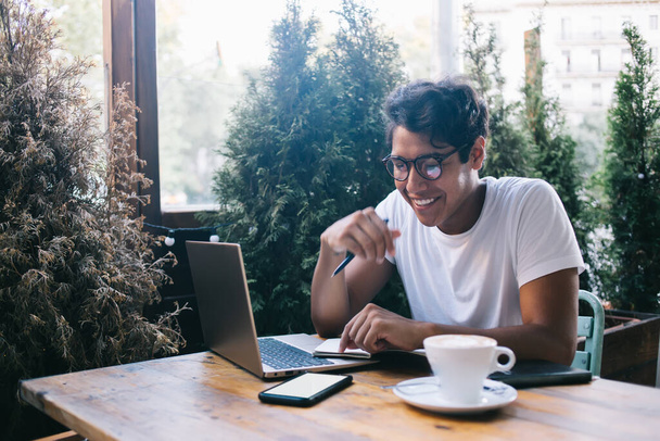 Cheerful hipster guy in optical eyewear enjoying freelance lifestyle sitting at cafe table and reading funny message on mockup smartphone gadget - 写真・画像