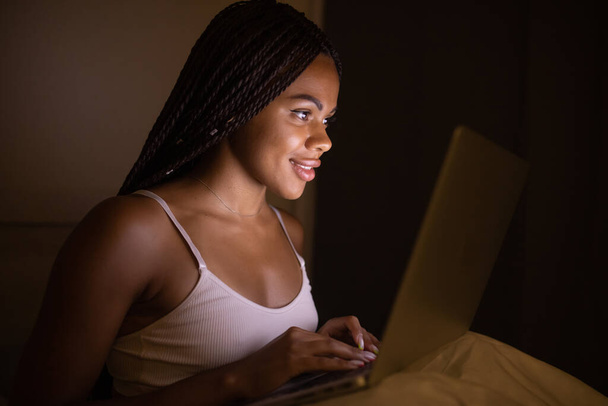 Happy pretty black woman using laptop on bed. Woman using laptop at night. - Фото, изображение