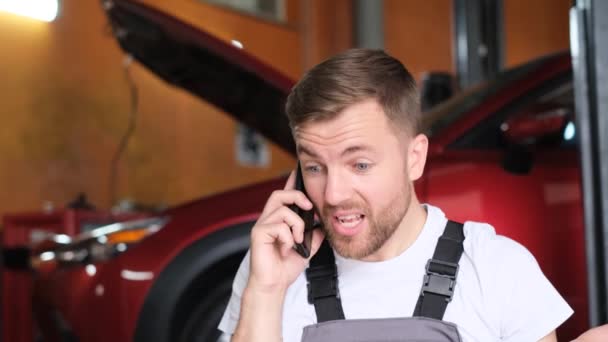 Egy dühös autószerelő beszél egy okostelefonon, hangosan kiabál a telefonba. Autószerelő a garázsban. Gépjármű-szolgáltatás - Felvétel, videó