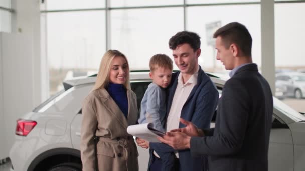 rodina v autosalonu, rodiče s mužským dítětem mluví s prodejcem aut o novém modelu auta v showroomu - Záběry, video