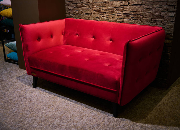 Стильный модерн ярко-красный мягкий велюровый диван, выставленный на продажу в мебельном дизайнерском магазине против стильной кирпичной стены в мебельном салоне интерьера освещенный мягким тусклым светом - Фото, изображение