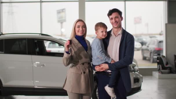 αγορά αυτοκινήτου, ευτυχισμένος άνθρωπος με αρσενικό παιδί στην αγκαλιά και τη γυναίκα κρατώντας τα κλειδιά για το νέο αυτοκίνητο, ενώ στέκεται στο κέντρο auto, χαμογελώντας και κοιτάζοντας κάμερα - Πλάνα, βίντεο