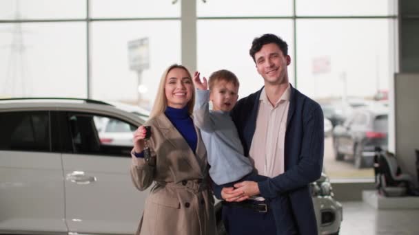 familia en auto show, cónyuges alegres con hijo en brazos sosteniendo las llaves de un automóvil nuevo mientras está de pie en el centro automático, sonriendo y mirando a la cámara - Imágenes, Vídeo