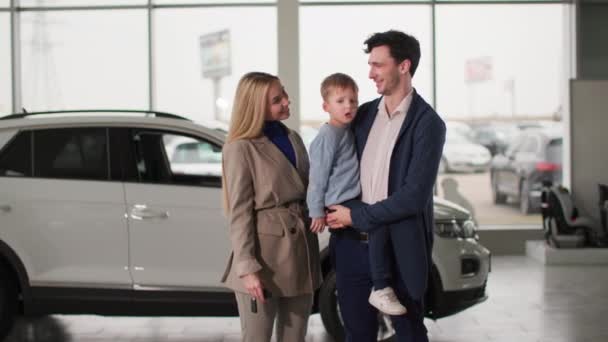 affari auto, sorridente uomo e donna con bambino in possesso di chiavi per il nuovo veicolo mentre in piedi nel centro auto, guardando la fotocamera - Filmati, video