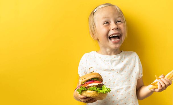 Cute little caucasian girl with blonde hair enjoying burger on a yellow background. Happy kid smiling and eating fast food burger - Foto, afbeelding