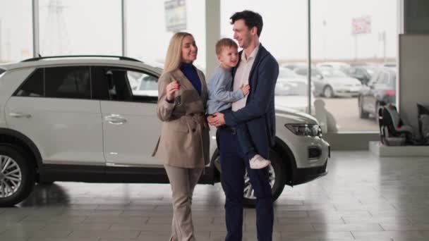 πορτρέτο της ευτυχούς γυναίκες και άνδρες αγοραστές με ένα μικρό αγόρι στην αγκαλιά τους κρατώντας τα κλειδιά για το νέο αυτοκίνητο στα χέρια, ενώ στέκεται στο κέντρο auto, χαμογελώντας και κοιτάζοντας κάμερα - Πλάνα, βίντεο