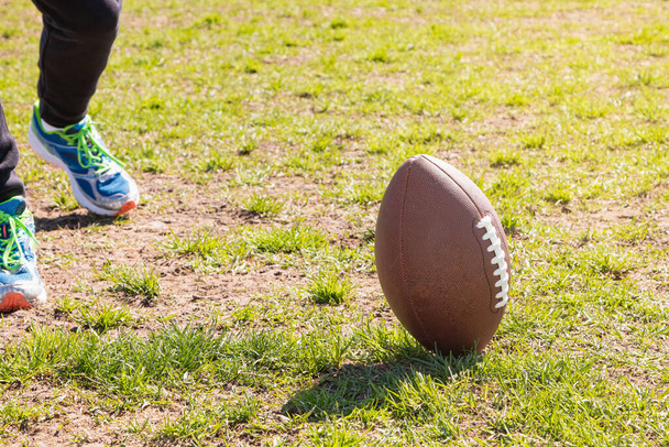 Em um campo de grama há uma bola de futebol americano e os pés de uma pessoa irreconhecível vestindo tênis. - Foto, Imagem