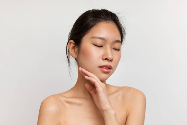 Señora coreana sensual desnuda tocando su cara y disfrutando del tacto sedoso después de usar la nueva crema nutritiva, posando sobre fondo de estudio blanco, espacio para copiar - Foto, Imagen
