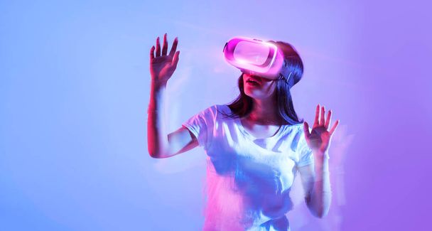 Σύγχρονες τεχνολογίες για διασκέδαση, κολάζ. Ενθουσιασμένη νεαρή γυναίκα που φοράει γυαλιά VR κοιτάζοντας χώρο αντιγραφής, βιώνει την εικονική πραγματικότητα, στέκεται σε μωβ φόντο σε νέον φωτισμό, πανόραμα - Φωτογραφία, εικόνα