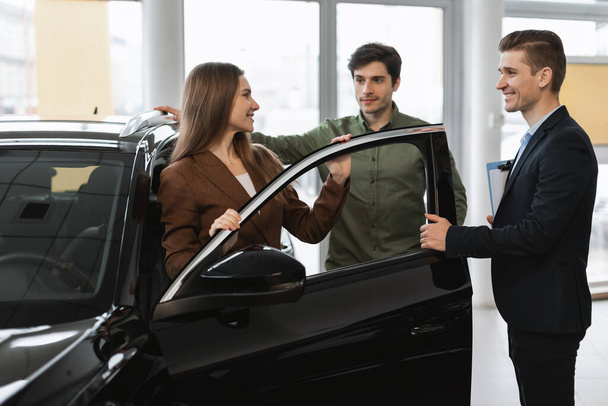 Positiivinen nuori pari keskustelee uuden auton ostamisesta tai vuokraamisesta automyyjän kanssa nykyaikaisessa jälleenmyyntisopimuksessa. Vuosituhannen valkoihoiset puolisot valitsevat auton, puhuvat showroom-johtajalle - Valokuva, kuva