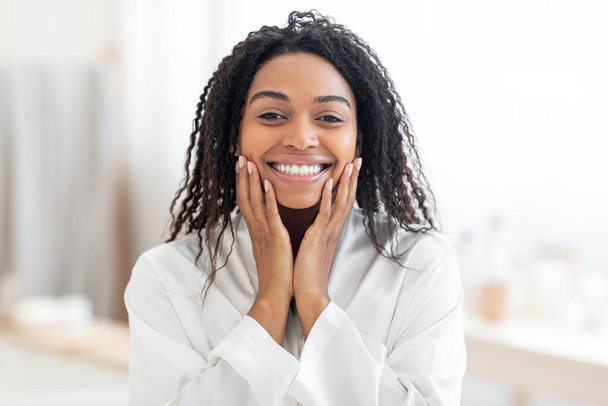Красивая молодая черная женщина трогает ее лицо и улыбается в камеру, делая косметические процедуры дома, привлекательная африканская американская женщина в белом шелковом халате наслаждаясь самообслуживания, копировать пространство - Фото, изображение