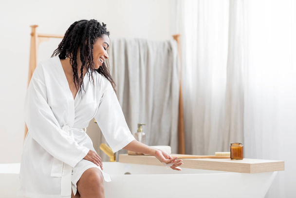 Home Spa. Belle femme noire assise sur la baignoire et la mousse touchante, attrayante jeune femme afro-américaine portant une robe de soie blanche se préparant à prendre un bain, se détendre dans la salle de bain à la maison, Copier l'espace - Photo, image