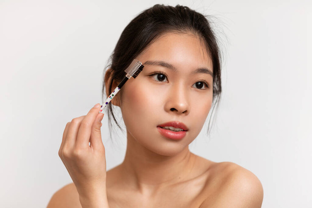 Natural makeup concept. Young korean lady brushing eyebrows, holding brush and doing morning beauty routine, standing over white background. Female cosmetics product - Фото, изображение