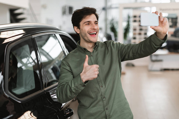 Aufgeregter junger Kaukasier macht ein Selfie mit dem Handy und zeigt Daumen hoch in der Nähe seines neuen Autos in einem modernen Autohaus. Schöner Millennial-Mann empfiehlt coolen Autosalon - Foto, Bild