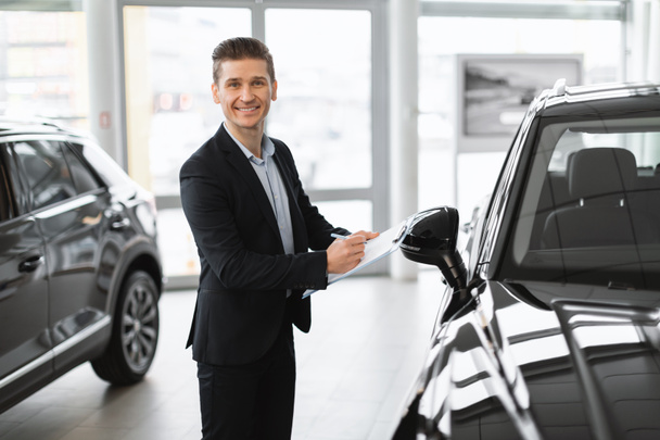 Positive young car salesman standing near new auto, taking notes and smiling at camera in automobile dealership, copy space. Millennial sales manager making vehicle checkup, working at showroom store - Foto, Imagen