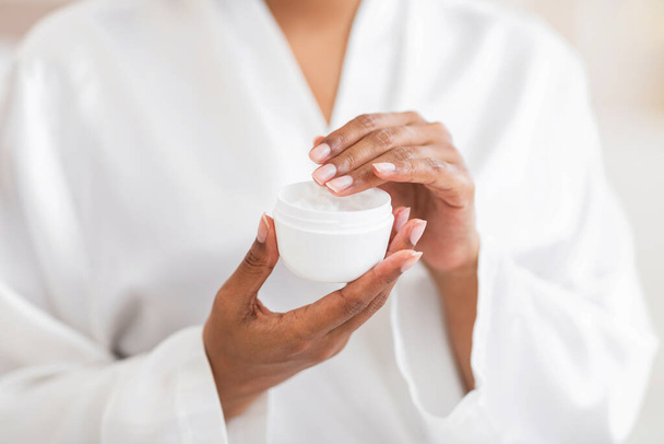 Closeup Shot Of Unrecognizable Black Woman Holding Opened Jar With Moisturising Cream, Young African American Female Wearing White Silk Robe Making Beauty Skincare Routine At Home, Cropped - Foto, afbeelding