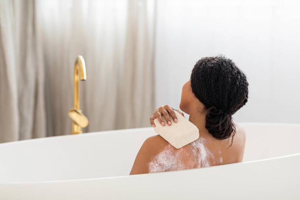 Nuori musta nainen pesee itsensä sienellä kylvyssä kotona, takana näkymä Afrikkalainen amerikkalainen nainen istuu kylpyammeessa vaahto ja hankausta iho, nauttien itsehoito rutiini, Kopioi tilaa - Valokuva, kuva