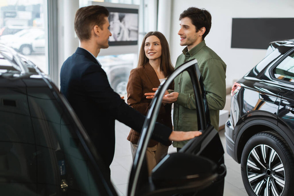 Χαρούμενος νεαρό ζευγάρι διαβούλευση με τον πωλητή αυτοκινήτων, επιλέγοντας νέο αυτοκίνητο στην αντιπροσωπεία αυτοκινήτων. Θετικοί Καυκάσιοι πελάτες επιλέγουν όχημα, μιλώντας με το διευθυντή στο showroom store - Φωτογραφία, εικόνα