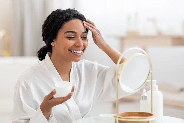 Красота. Привлекательная черная женщина, нанося увлажняющий крем на лицо, глядя в увеличительное зеркало в ванной комнате, красивая афро-американская женщина питательная кожа и улыбаясь отражению - Фото, изображение