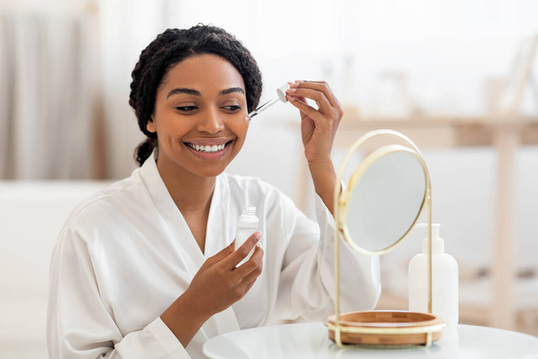 Concepto de cuidado de la piel. Hermosa dama negra aplicando suero facial con gotero mientras está sentada frente al espejo en casa, sonriendo mujer afroamericana hidratando la piel, disfrutando de la rutina de autocuidado - Foto, Imagen