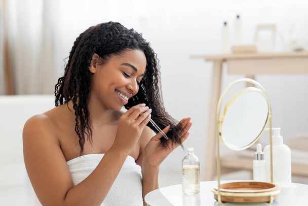 Onnellinen musta nainen soveltamalla kosteuttava öljy vaurioitunut hiukset päättyy suihkun jälkeen, hymyilevä Afrikkalainen amerikkalainen nainen istuu lähellä peiliä kylpyhuoneessa sisustus, joten hiustenhoito rutiini kotona, lähikuva - Valokuva, kuva