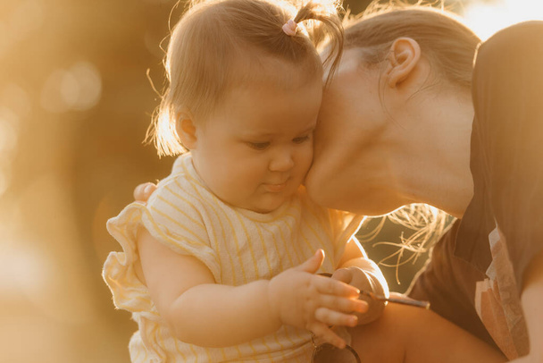 Uma criança feminina de roupas amarelas está brincando com óculos de sol perto de sua mãe nos raios de sol. Mãe está beijando sua filha no parque ao pôr do sol. - Foto, Imagem
