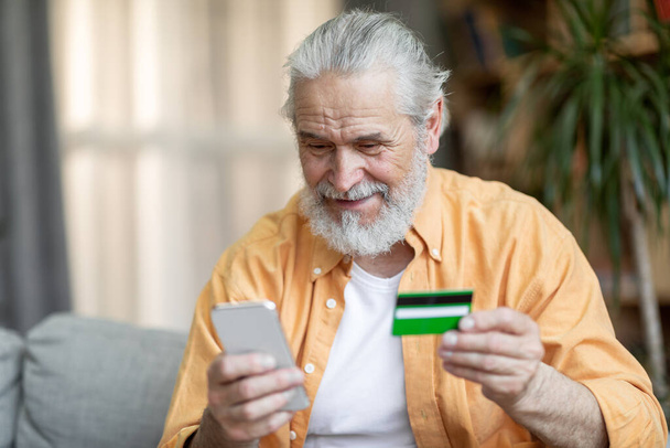 Ευτυχισμένος όμορφος γενειοφόρος ηλικιωμένος άνδρας κάθεται στον καναπέ, χρησιμοποιώντας σύγχρονο smartphone και πιστωτική κάρτα, ψώνια στο Διαδίκτυο, ενώ διαμένουν στο σπίτι, πληρώνουν για αγαθά και sevises σε απευθείας σύνδεση, αντίγραφο χώρου - Φωτογραφία, εικόνα