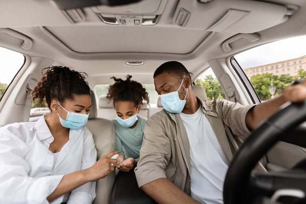 Arabada ellerinde dezenfektan spreyi olan siyah bir aile. Ebeveynler ve kızları otomobille seyahat ediyorlar, yüz maskeleri takıyorlar ve 19 numaralı koruma için ellerini dezenfekte ediyorlar. - Fotoğraf, Görsel