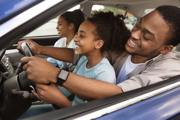 Cheerful African American Dad Teaching Preteen Daughter To Drive A Car. Kid Sitting On Fathers Knees Driving Together And Having Fun While Family Traveling By Automobile. Selective Focus - Photo, image
