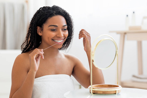 Оральный уход. Улыбающаяся черная женщина, смотрящая в зеркало и пользуясь зубным налетом в ванной, привлекательная молодая афроамериканка, очищающая зубы, делающая утреннюю гигиену дома, крупный план - Фото, изображение
