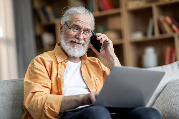 Positiivinen komea vanhusten mies, jolla on parta istuu sohvalla, käyttää nykyaikaista kannettavaa tietokonetta ja ottaa puhelimen keskustelun perheen kanssa, pyytää apua, kun käytät tietokonetta kotona, kopioi tilaa - Valokuva, kuva